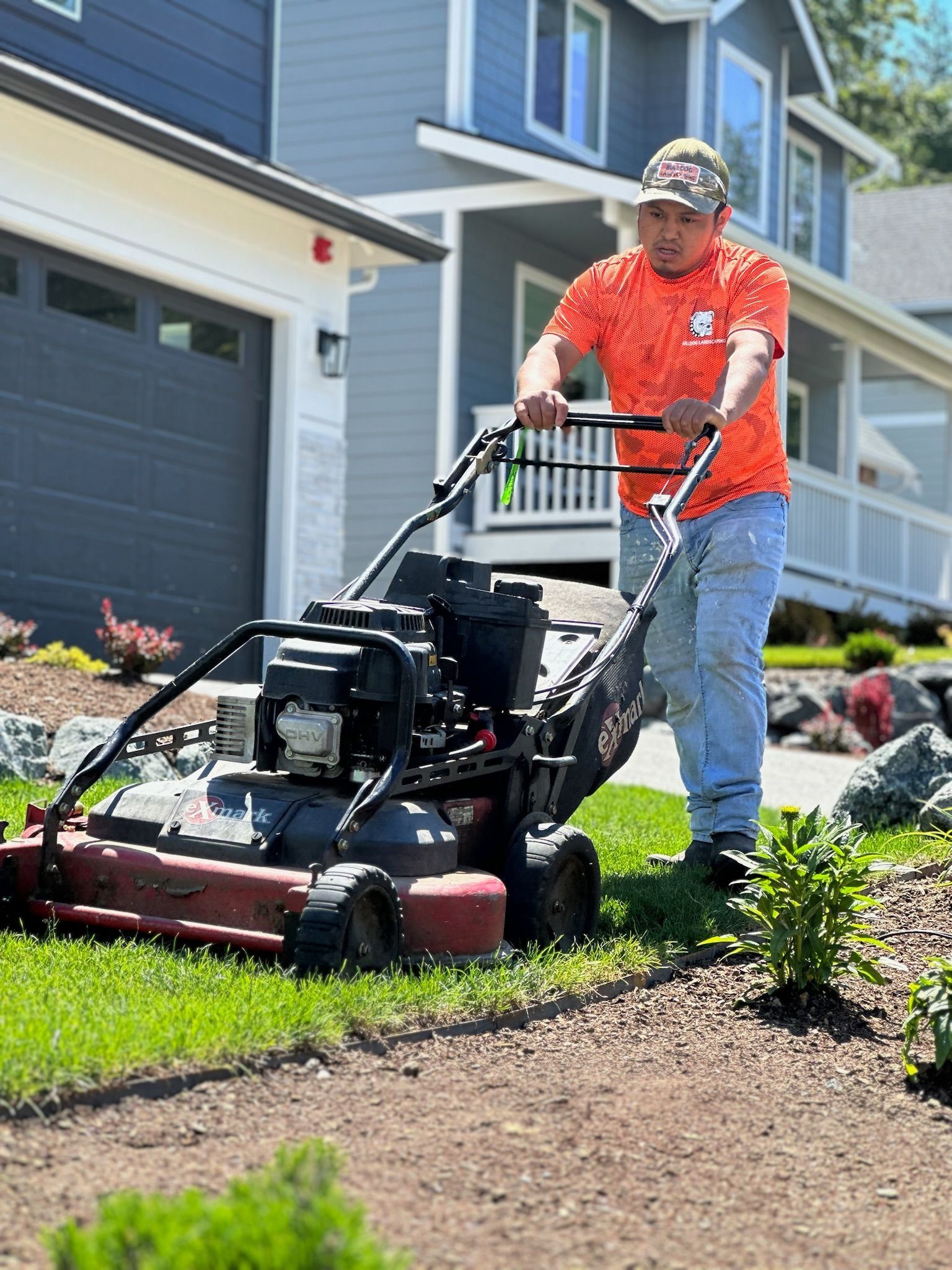 Person mowing a lawn in front of a house. He is wearing an orange shirt and jeans.