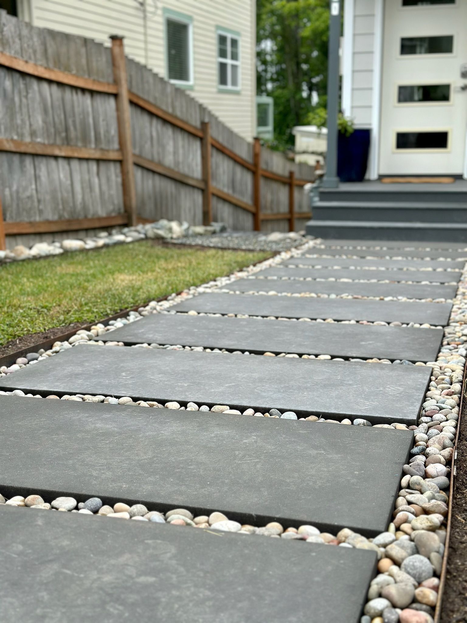 Gray stepping stones lead to a white door, bordered by gravel and grass. A wooden fence is in the background.
