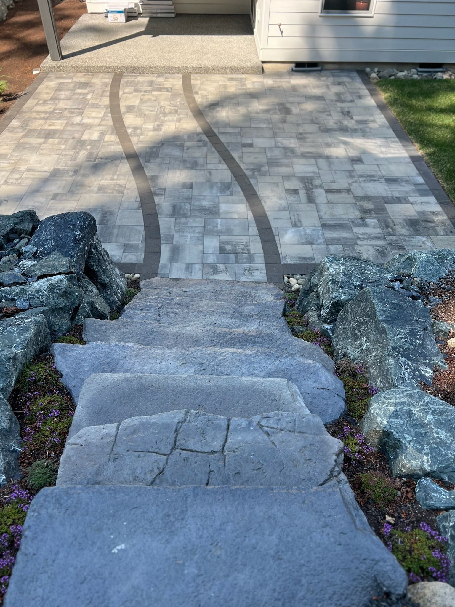 Stone steps leading up to a paved walkway with a curved dark border, bordered by large rocks.