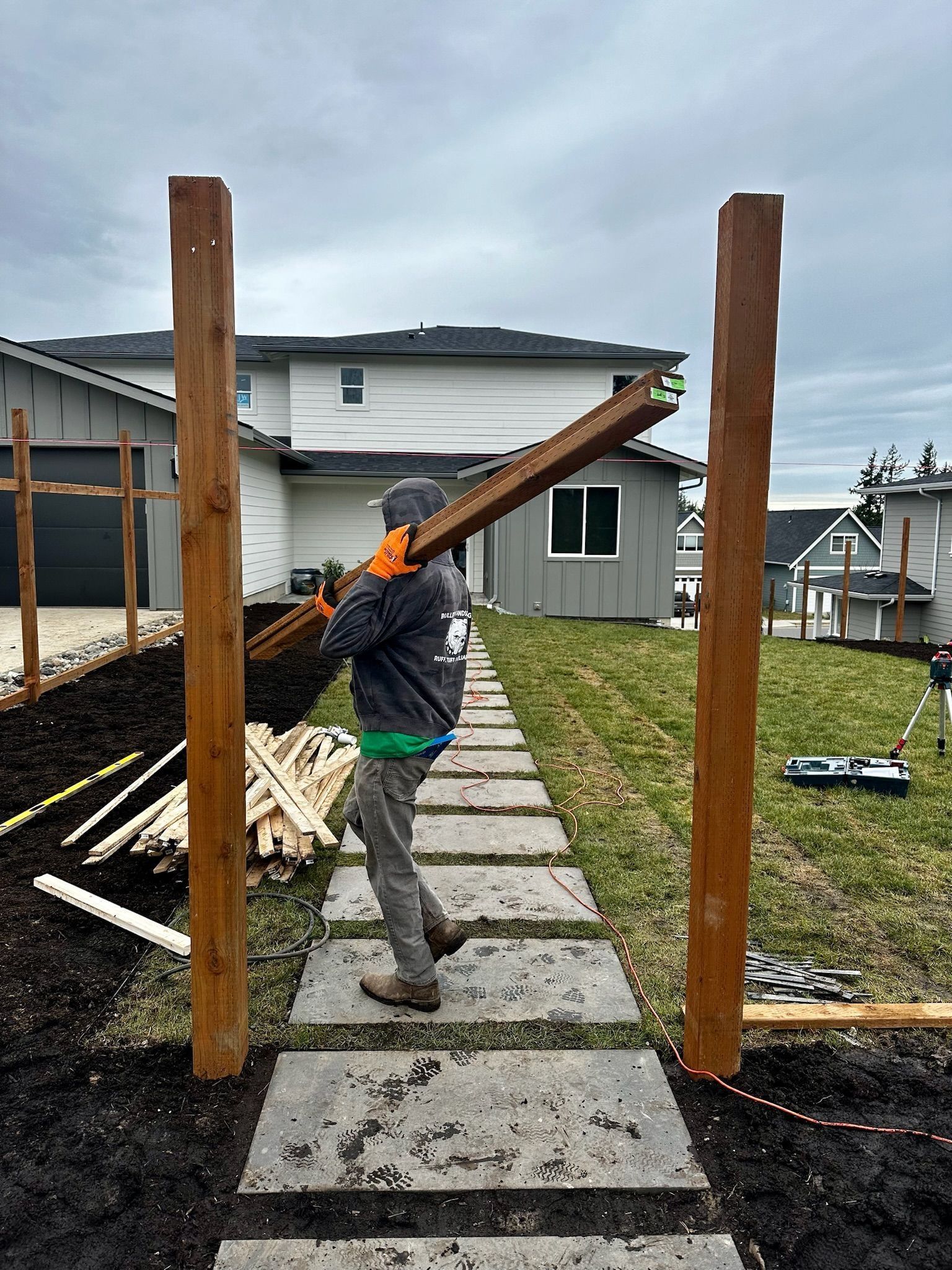 Man carrying wood plank between two fence posts on a stone path.