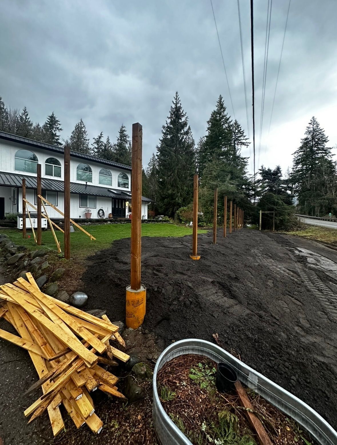 Fence posts installed in dark soil in front of a white house with trees in the background.