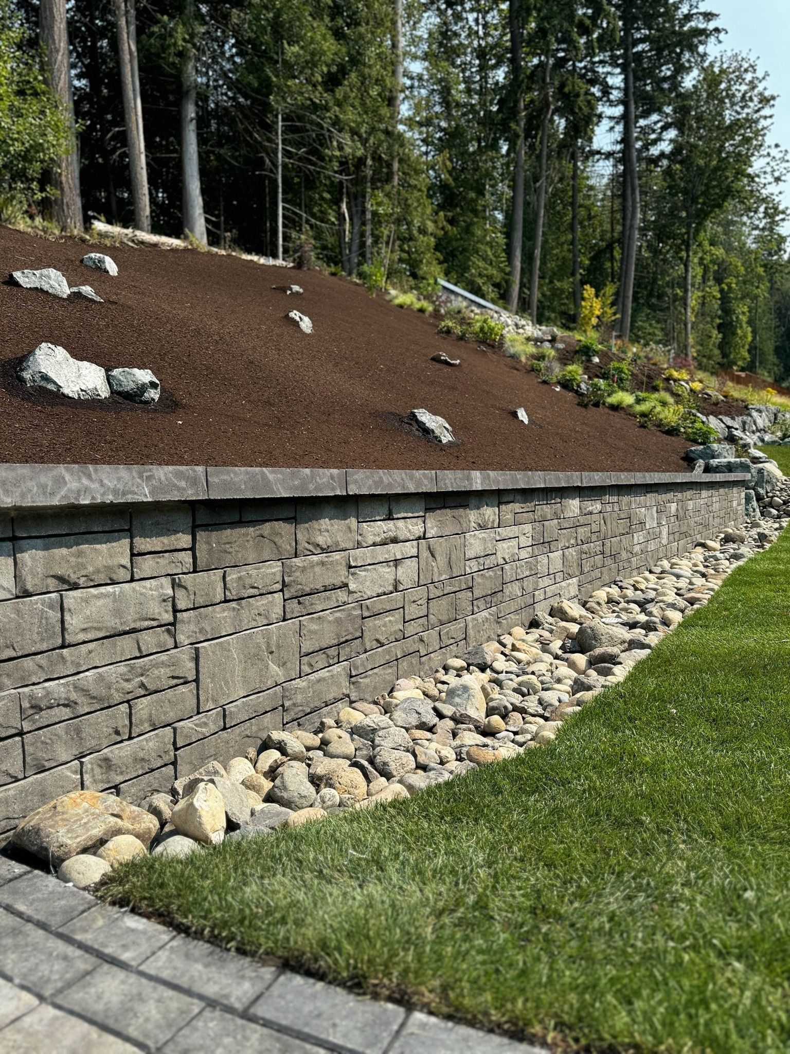 Stone retaining wall supporting a hillside with mulch, small rocks, and green grass.