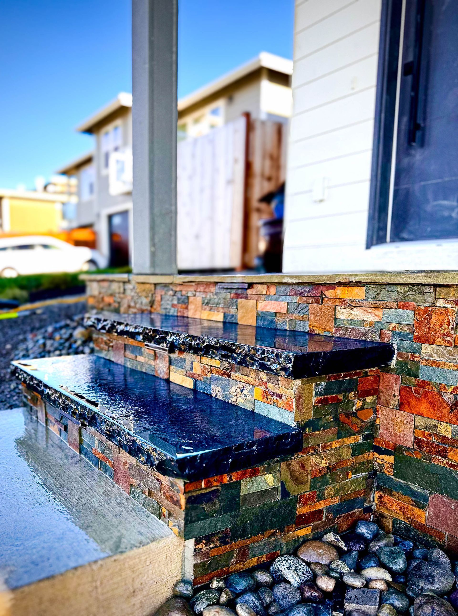 Stone steps leading to a house with layered colorful brickwork, wet from rain, and small river rocks.