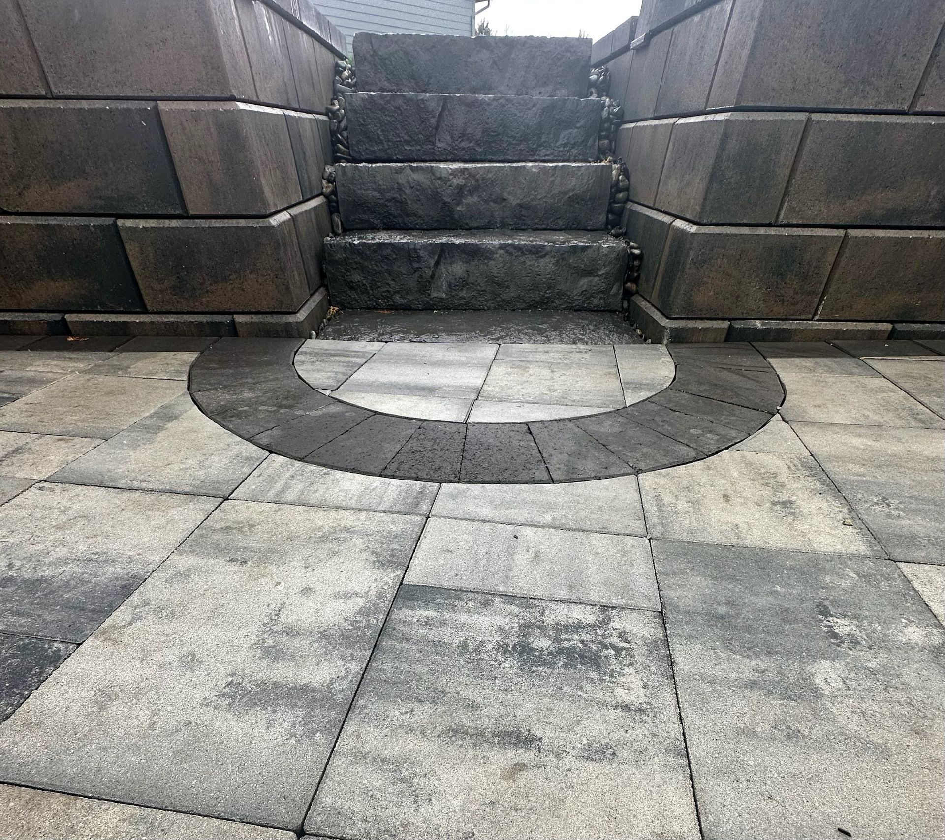Stone steps leading up, framed by dark stone blocks. Circular dark stone inlay on light stone patio.