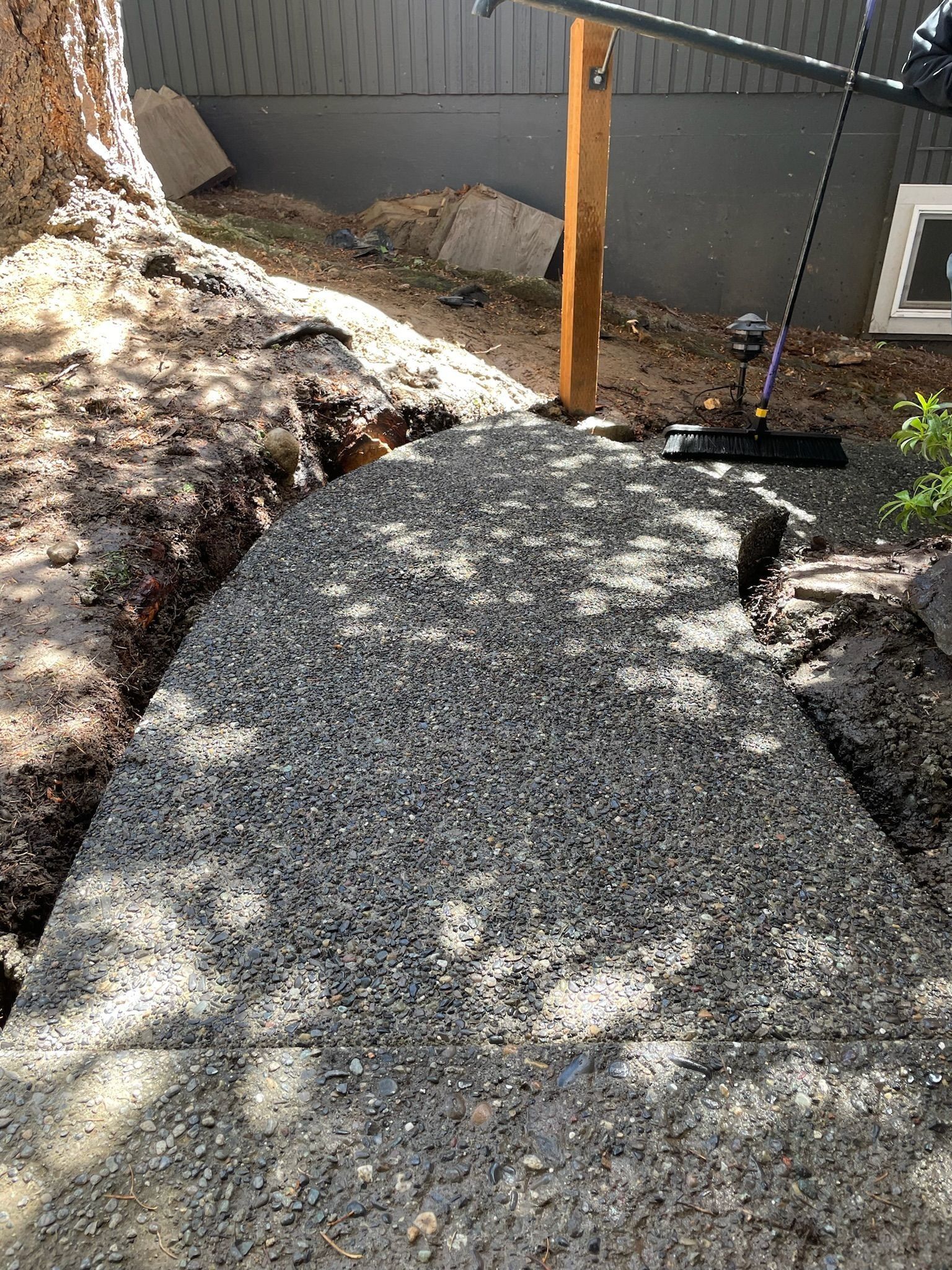 Pathway of gravel in a dirt trench, near a tree and building. A broom is propped nearby.