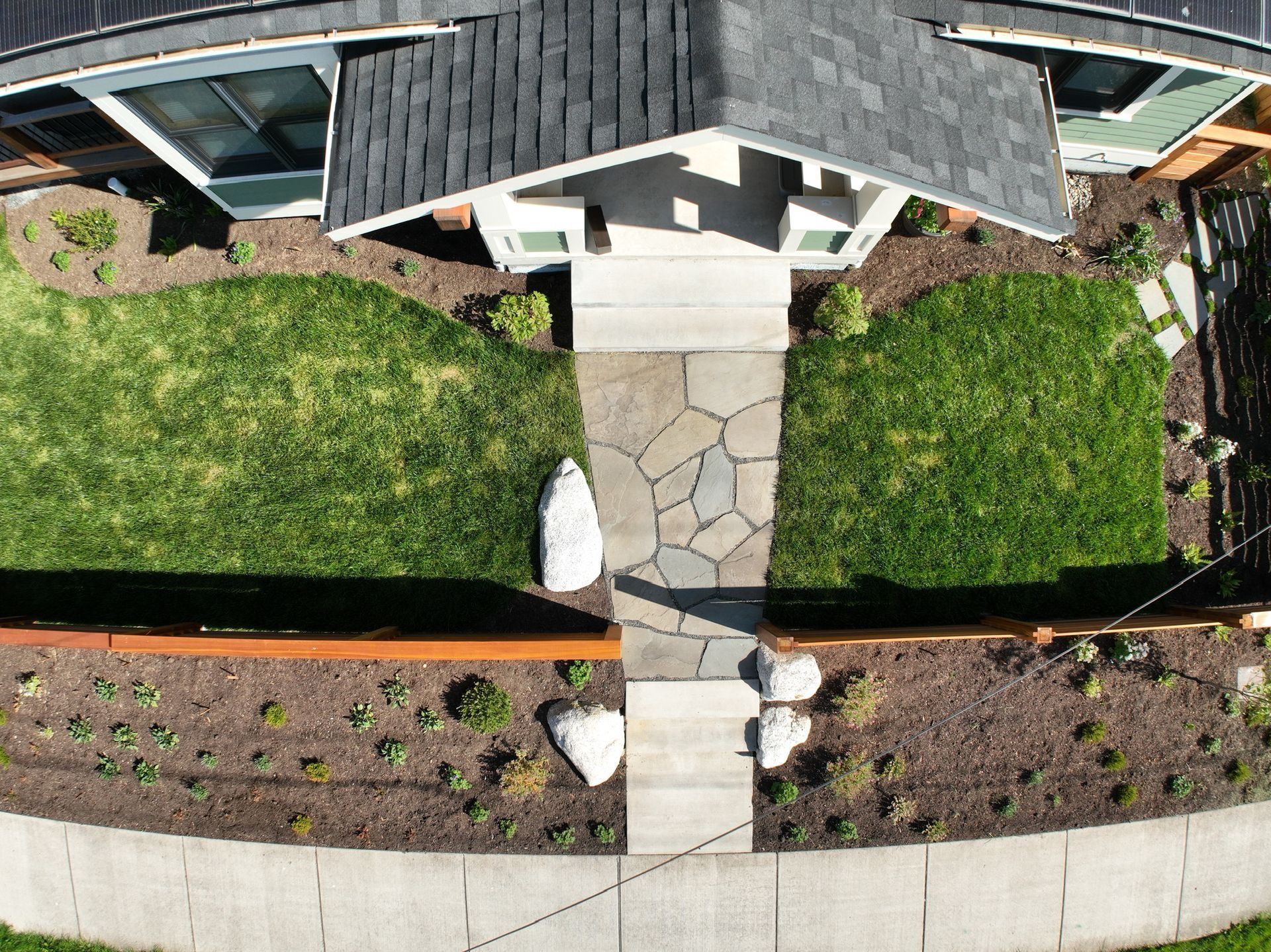 Overhead view of a house with a stone pathway, flanked by green lawns and brown flowerbeds, leading to the entrance.