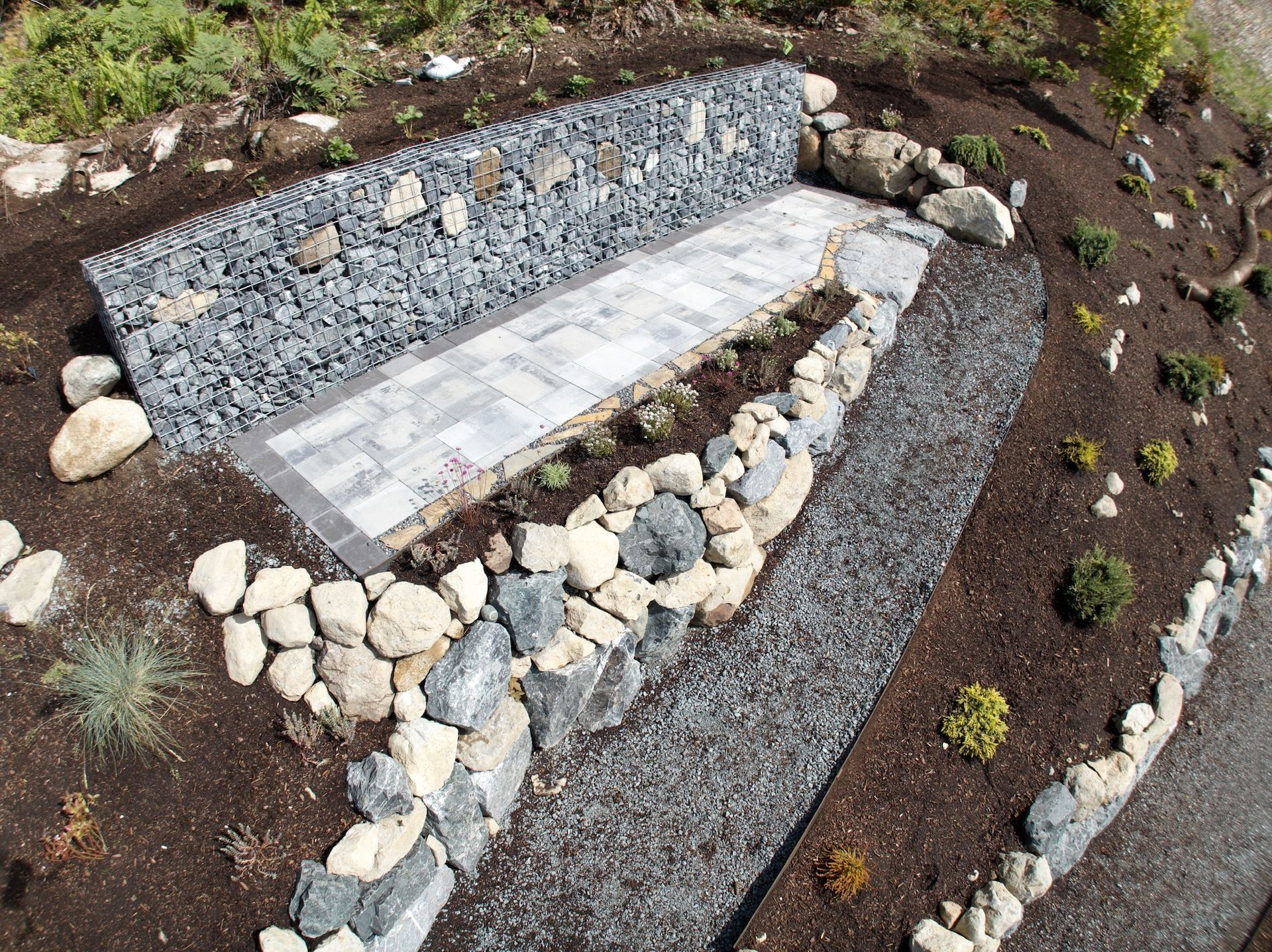 Stone and gabion planter with a paved walkway, surrounded by gravel and mulch.
