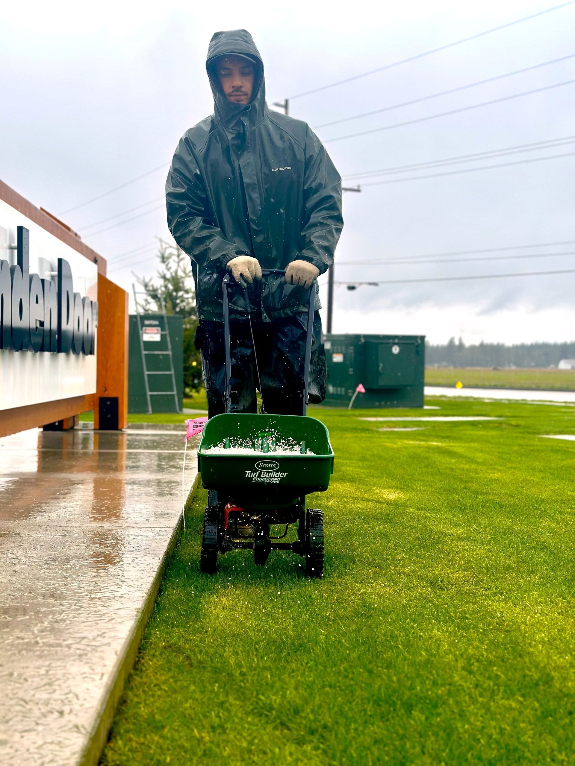 Man in rain gear pushing a spreader on a wet lawn.
