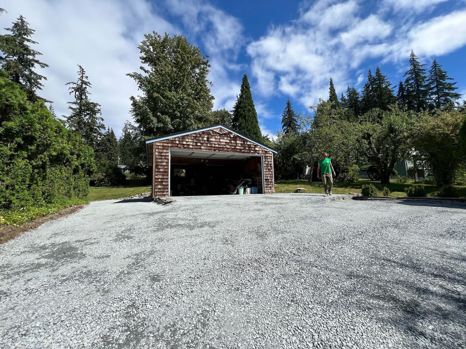 Gravel driveway leading to an open garage with brick facade, trees in the background, blue sky with clouds.