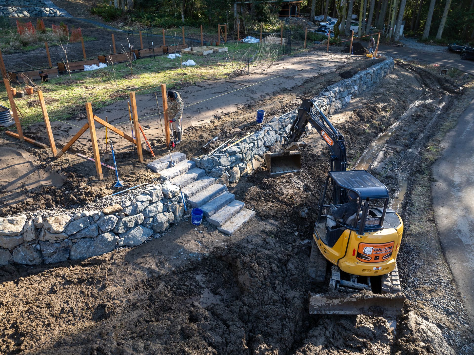 Yellow excavator digging earth beside a stone retaining wall at a wooded construction site
