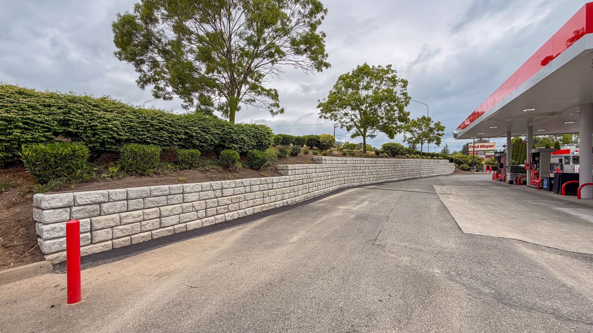 Gas station driveway beside a stone retaining wall and trees, with a red roof canopy on the right