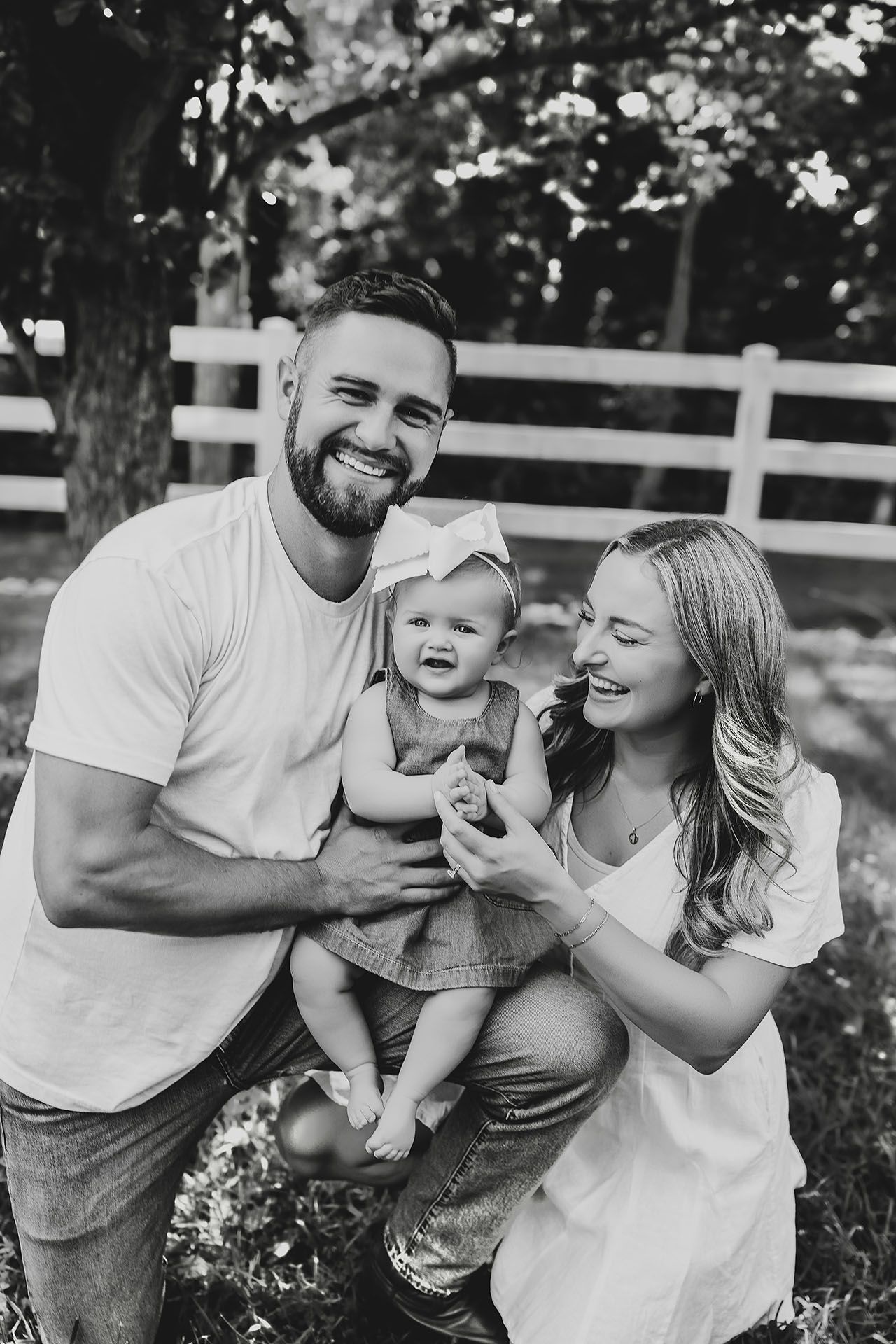 Family portrait in black and white: Father, mother, and baby smiling outdoors. White fence background.