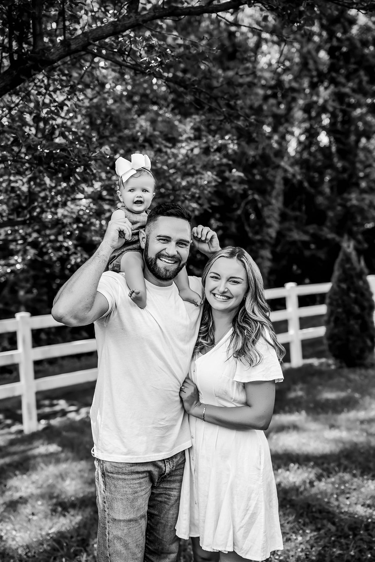 Family of three poses outdoors; father holds child on shoulders, mother smiles. Black and white.