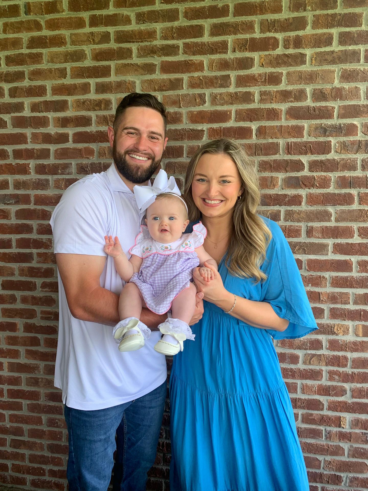 A family of three smiling in front of a brick wall. The parents hold a baby girl.