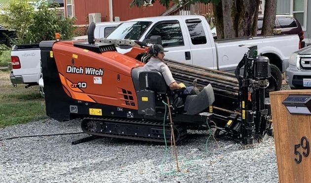 A worker operates an orange Ditch Witch JT10 horizontal directional drill on a gravel lot near parked vehicles.