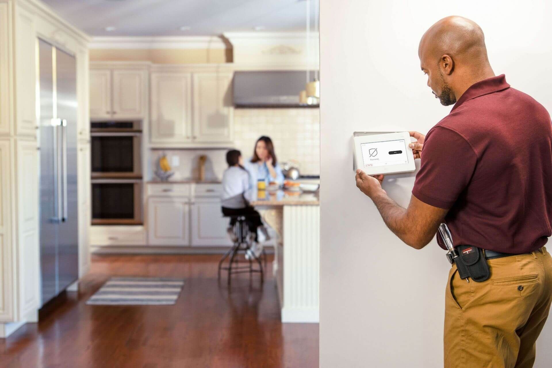 A man in a burgundy shirt adjusts a wall-mounted control panel; a kitchen with a woman and child is in the background.