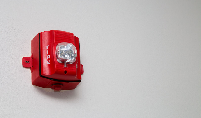 Red fire alarm with a strobe light on a white wall.