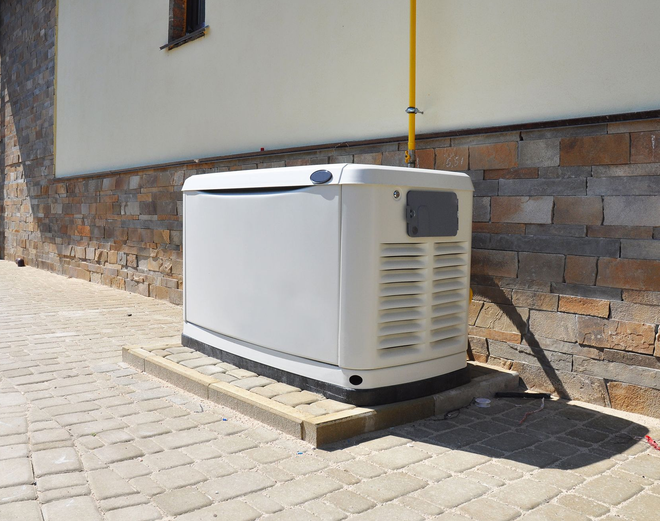 A white generator on a brick base next to a house with a brick wall and a yellow gas line.