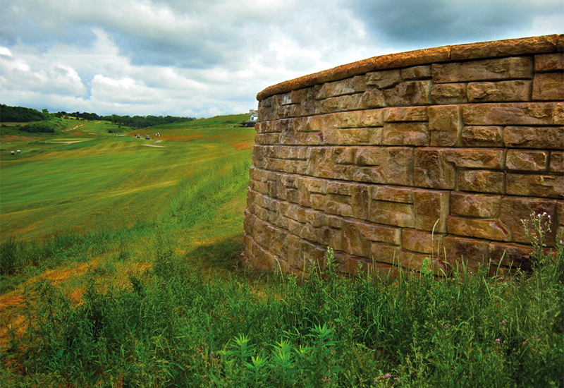 A large stone wall surrounds a grassy field with a golf course in the background