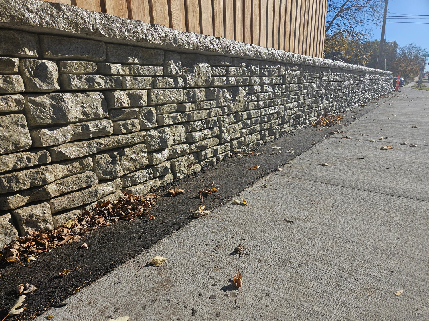 A stone wall along a sidewalk next to a wooden fence.