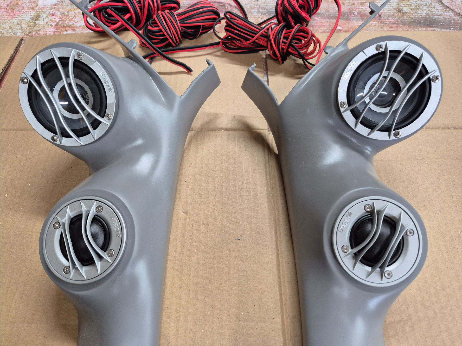 Two gray car speaker pods with metal grilles and wiring on a brown surface.