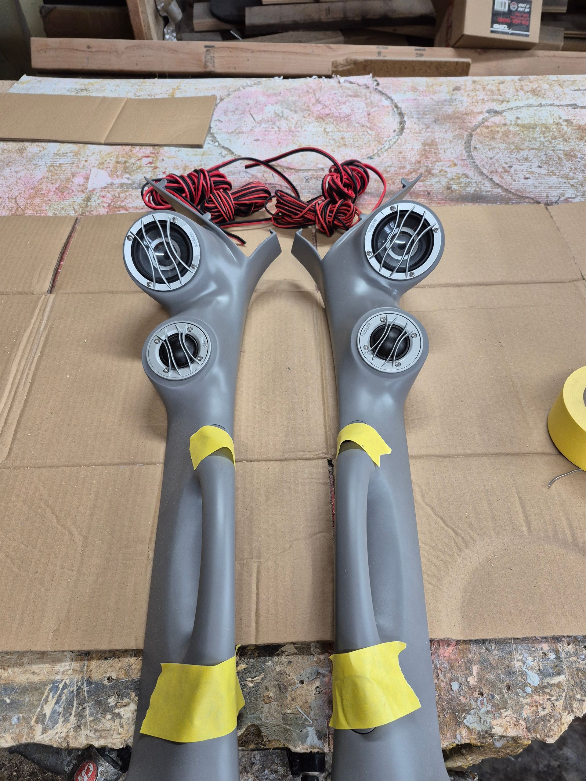 Two gray automotive pillar speaker pods with speakers installed, on a workbench.