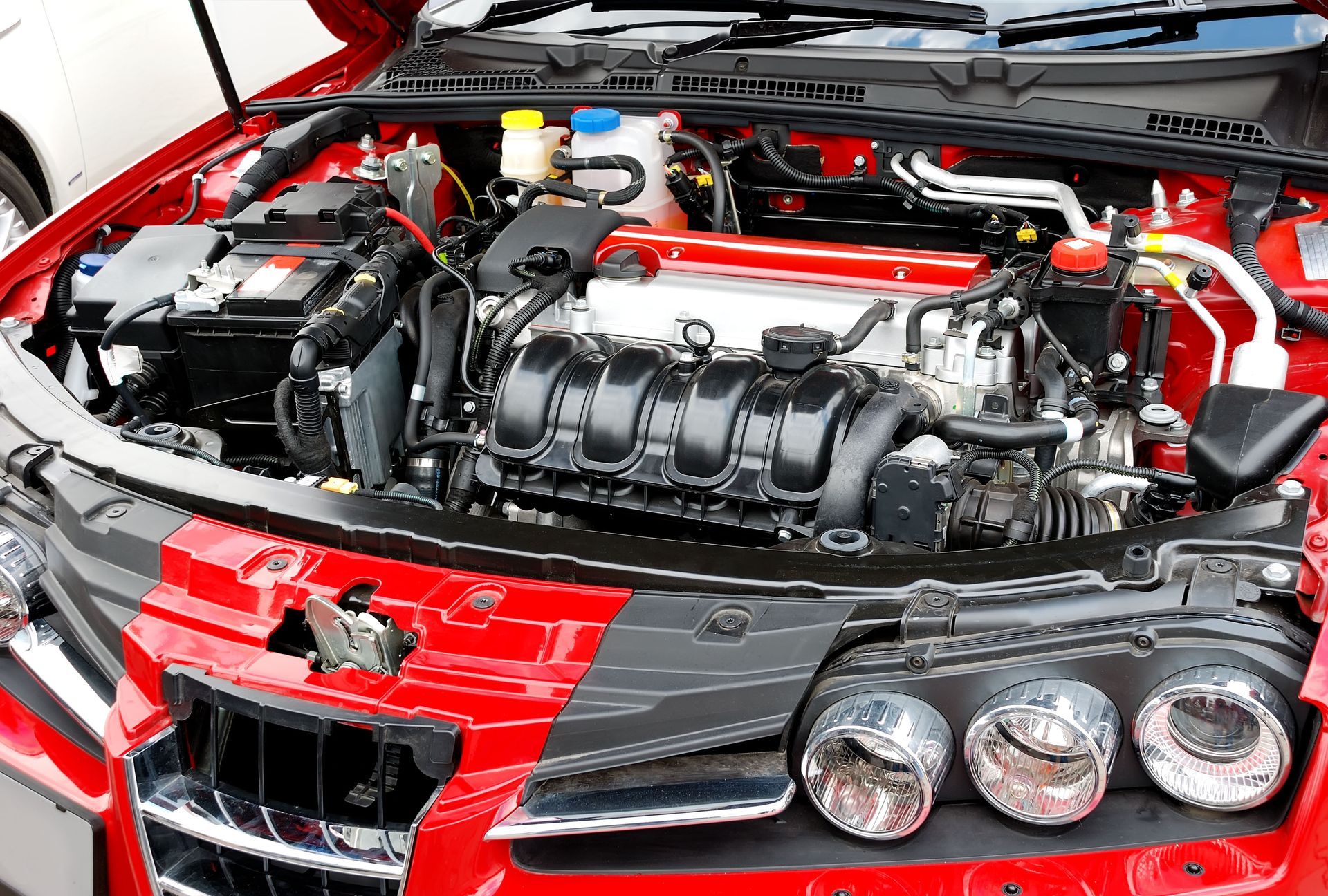 Close-up of a red sports car engine bay with black intake components and three round headlights visible.