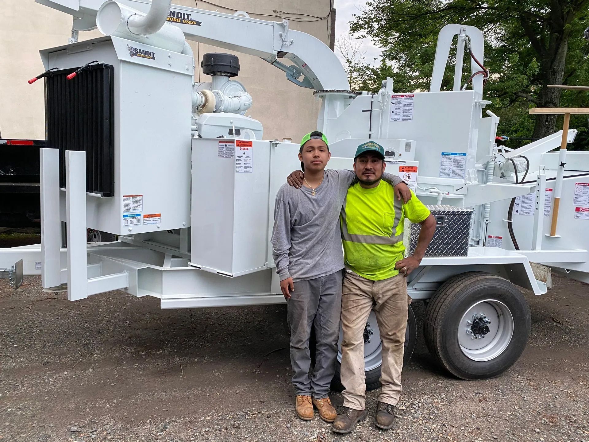 Two men stand by a large white wood chipper outdoors. One man wears a neon green vest and has his arm around the other.