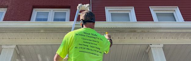 A person in a neon green shirt cleans gutters with a pole in front of a red-roofed building.