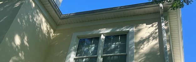 Exterior view of a cream-colored building with a window; shadows from tree branches fall across the wall and the clear blue sky.
