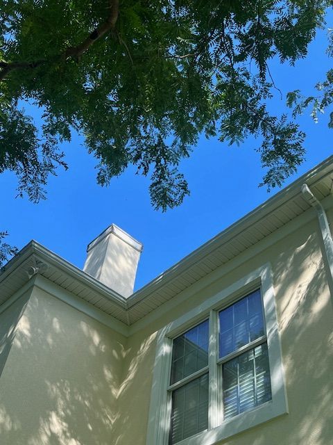 Beige house exterior with chimney, gutters, and windows against a blue sky, shaded by tree branches.