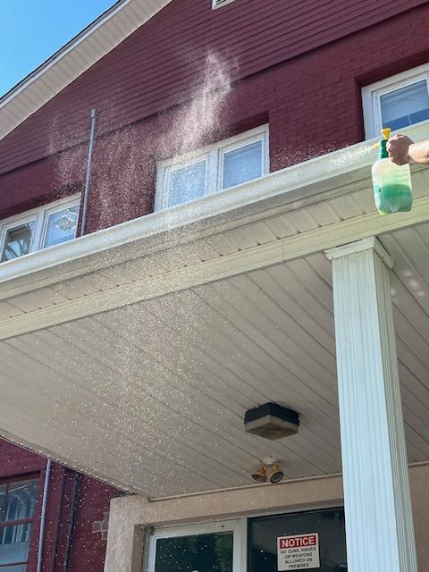 Person spraying a red brick building's gutters with a green liquid from a spray bottle.