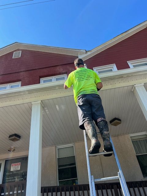 Man on ladder power washing a house's trim. He wears a neon shirt, camo boots. Red building, blue sky.
