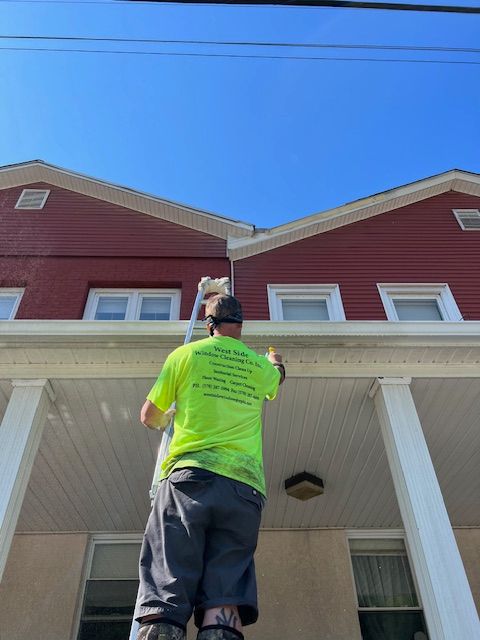 Person painting trim on a house from a ladder, with a blue sky overhead. The house has red siding.