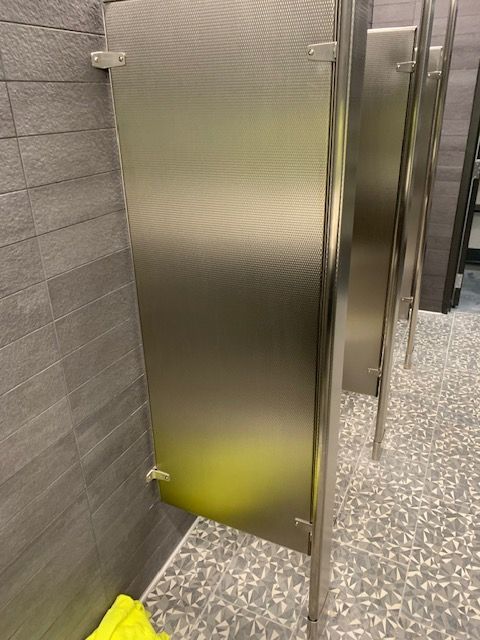 Stainless steel public restroom stalls in a row, with a gray tiled wall and speckled floor.