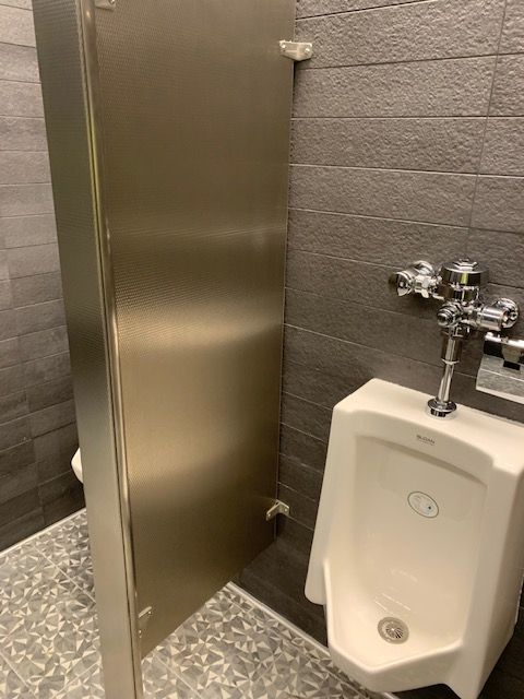 Urinal and stall in a public restroom. Stainless steel stall door, white urinal, gray tiled walls.