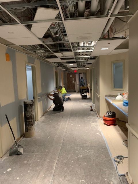 Construction workers in hallway, installing drywall. Exposed ceiling, debris on floor.