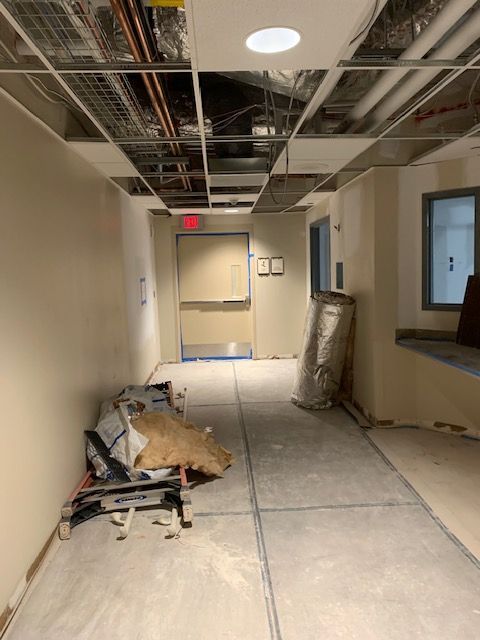 Hallway under construction, unfinished walls, ceiling framework, concrete floor, exit sign at the end.