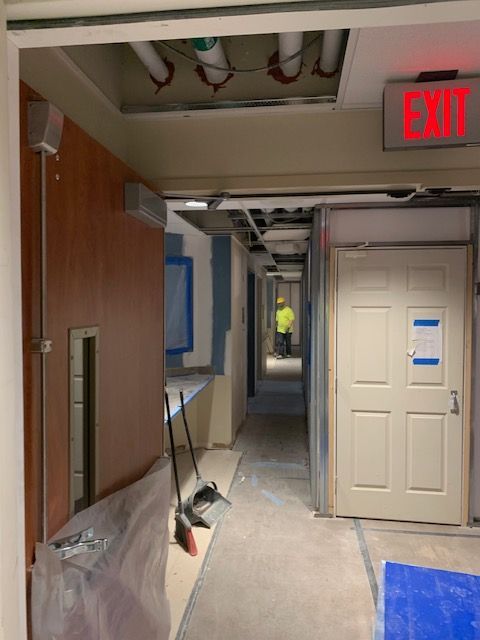 Interior hallway under construction; a worker in safety vest visible at the end. Exit sign.