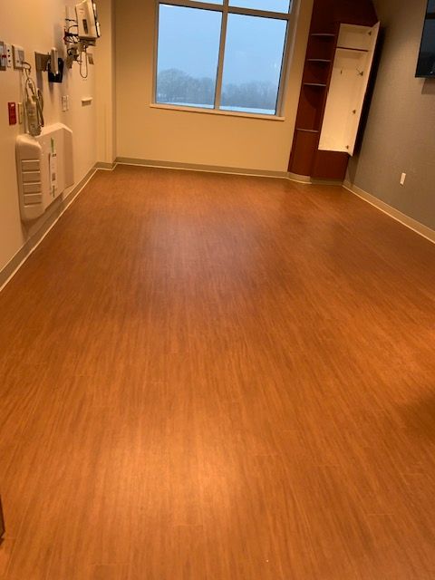 Empty medical room with wood-look flooring, large window, medical equipment on wall.