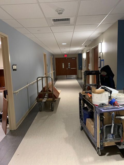 Hospital hallway with blue walls, open doors, and carts. An exit sign is visible at the end.