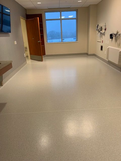 Empty medical exam room with gray floor, tan walls, and a large window.