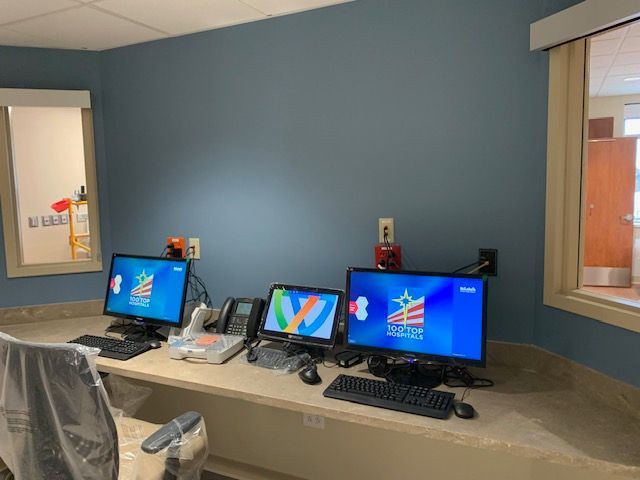 Three computer monitors on a desk in a blue-walled room; a window on either side of the desk.