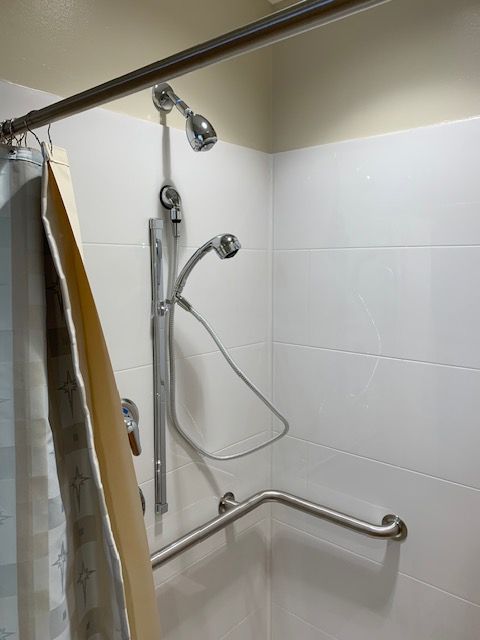 Shower with white tiled walls, grab bar, adjustable shower head, and curtain.