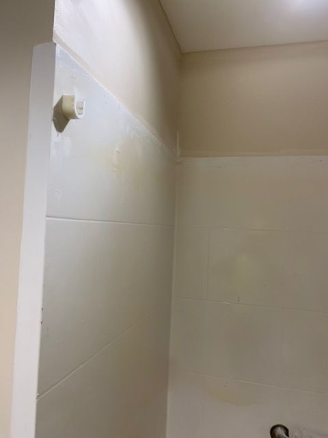 Corner of a tiled shower with a pipe, cream-colored walls, and a light source.