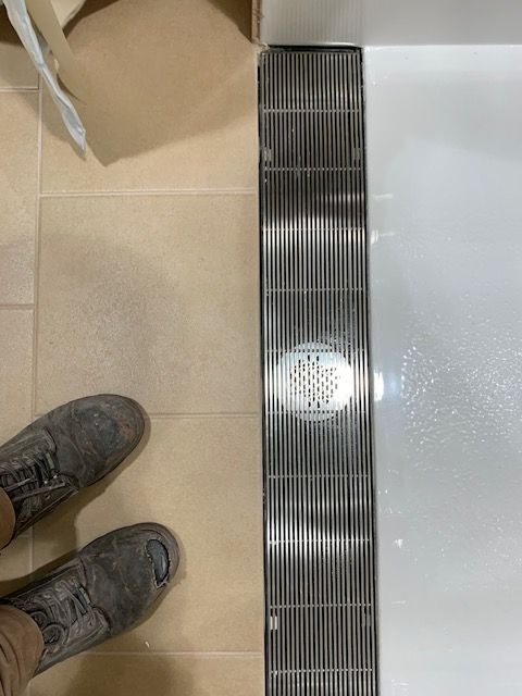 A metal shower drain in a bathroom. Shoes and beige tile are visible.