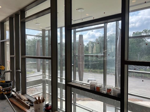 Interior view of a building with large windows and metal frames. Outside are trees and tall art installations.