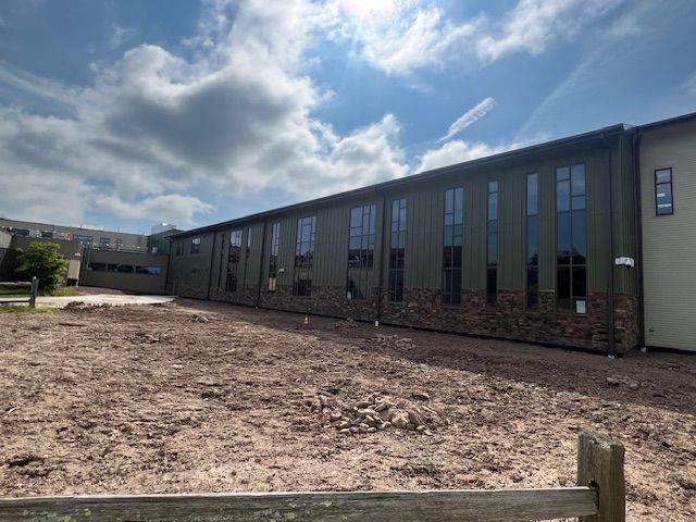 Building with green metal siding and tall windows, under construction, against a cloudy sky.