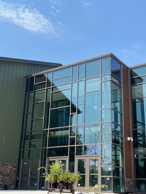 Modern building exterior with glass and dark frame. Green siding to the left, blue sky overhead.