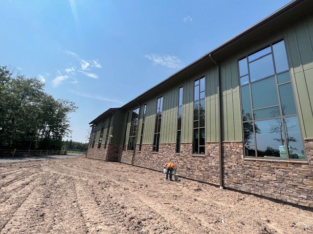Exterior view of a new building under construction, green siding, stone base, large windows, blue sky, worker in orange vest.